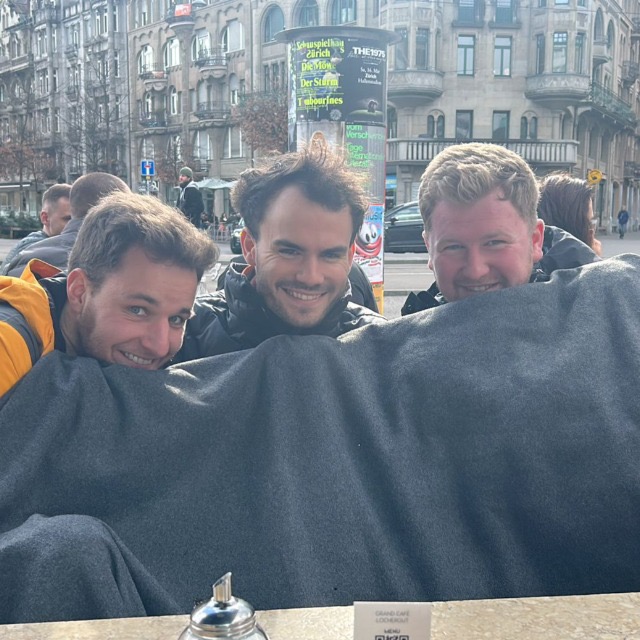 Three friends smiling at an outdoor café, sharing a blanket across their laps on a chilly day.